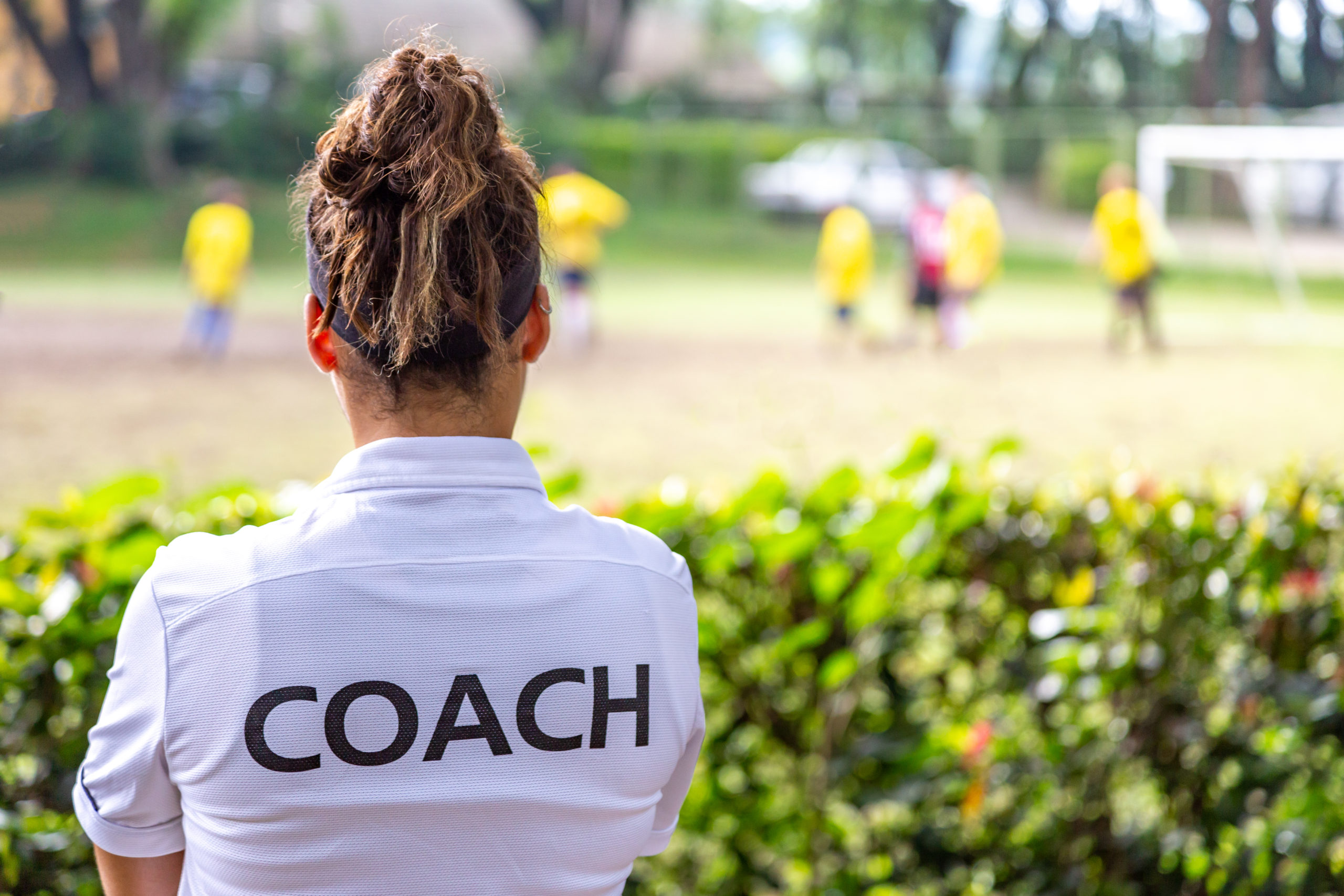 Female coach standing beside blurred playing field, her back to the camera with "Coach" written on her shirt.